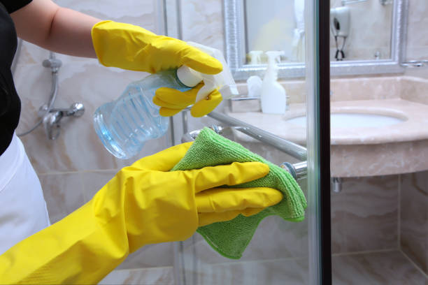 Person wearing yellow gloves uses spray bottle and cloth to clean bathroom mirror.